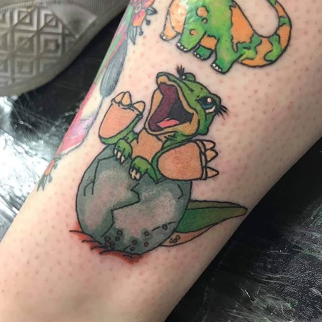 Cartoon tattoo of Ducky dinosaur in the egg on the left leg
