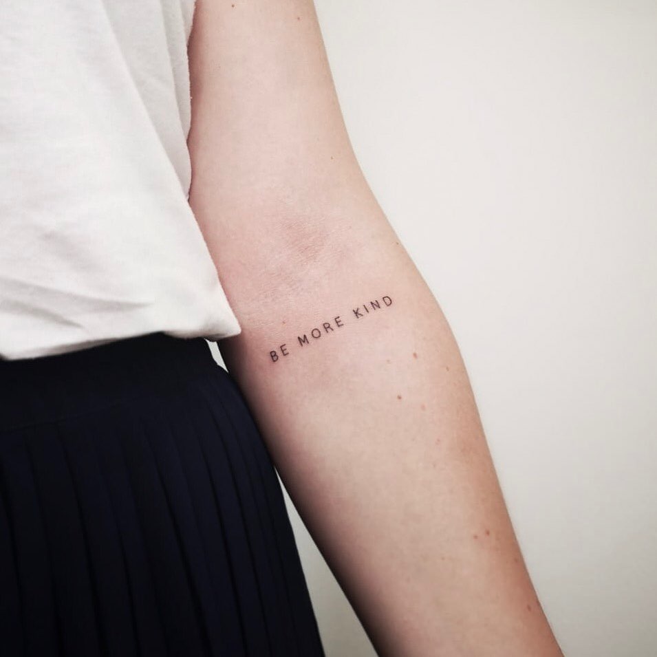Lettering black tattoo for women of "Be more kind" written on the left forearm